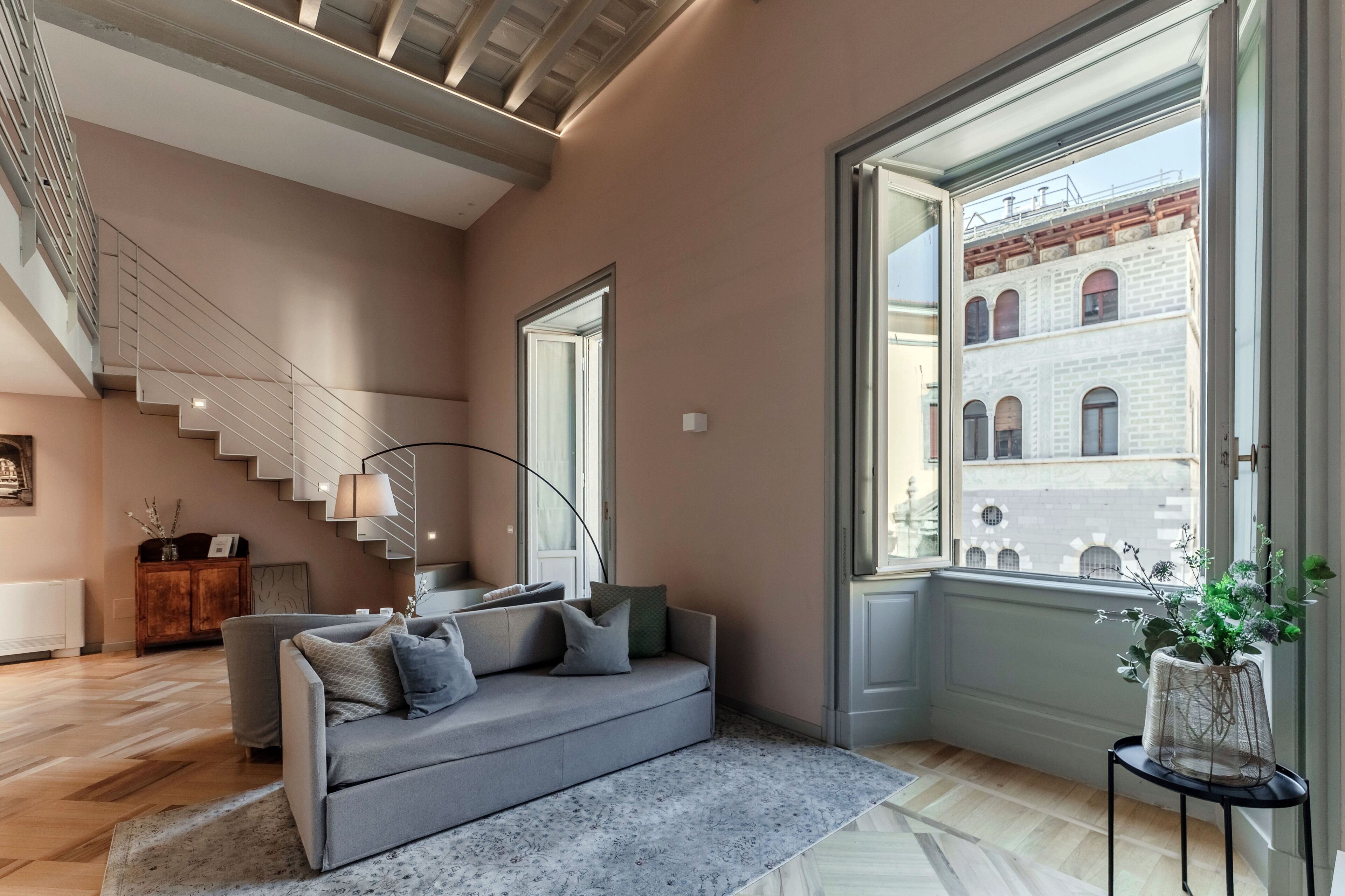 A modern living room with a staircase, a sofa, a coffee table, and a view of a building through a large window.