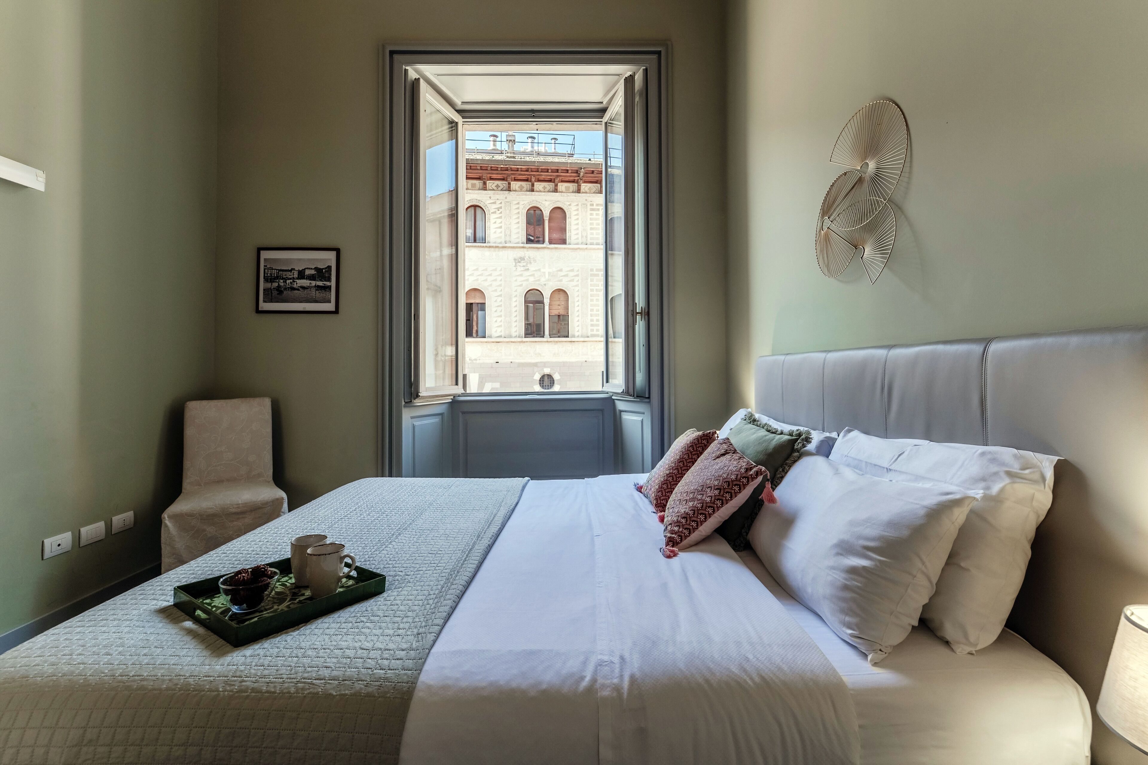 A hotel room with a bed, a window with a view of a building, a chair, a tray with a cup and a bowl, and a wall-mounted fan.