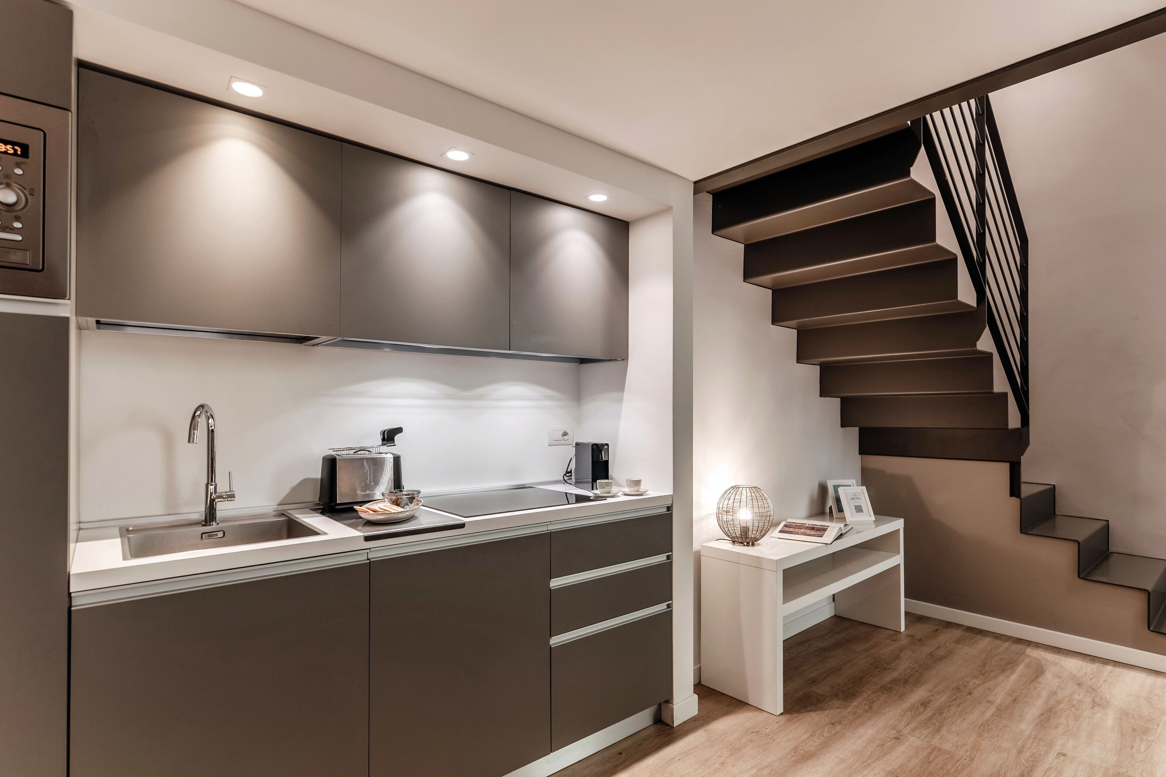A modern kitchen with a built-in stove, a sink, and a countertop. There is a staircase with a black railing leading to an upper level.