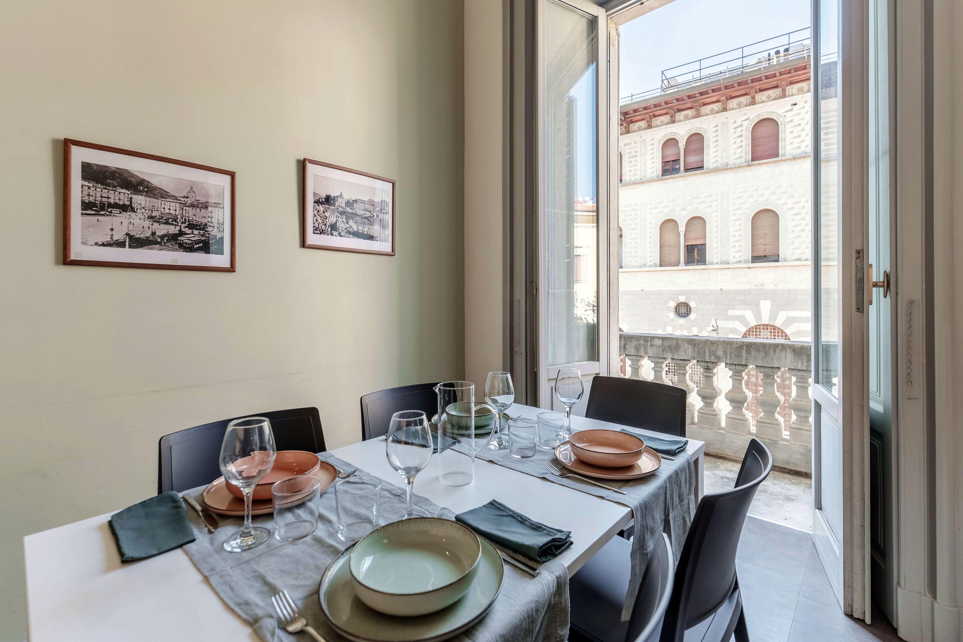 A dining table set with plates, glasses, and cutlery, a framed picture on the wall, and a view of a building through an open door.