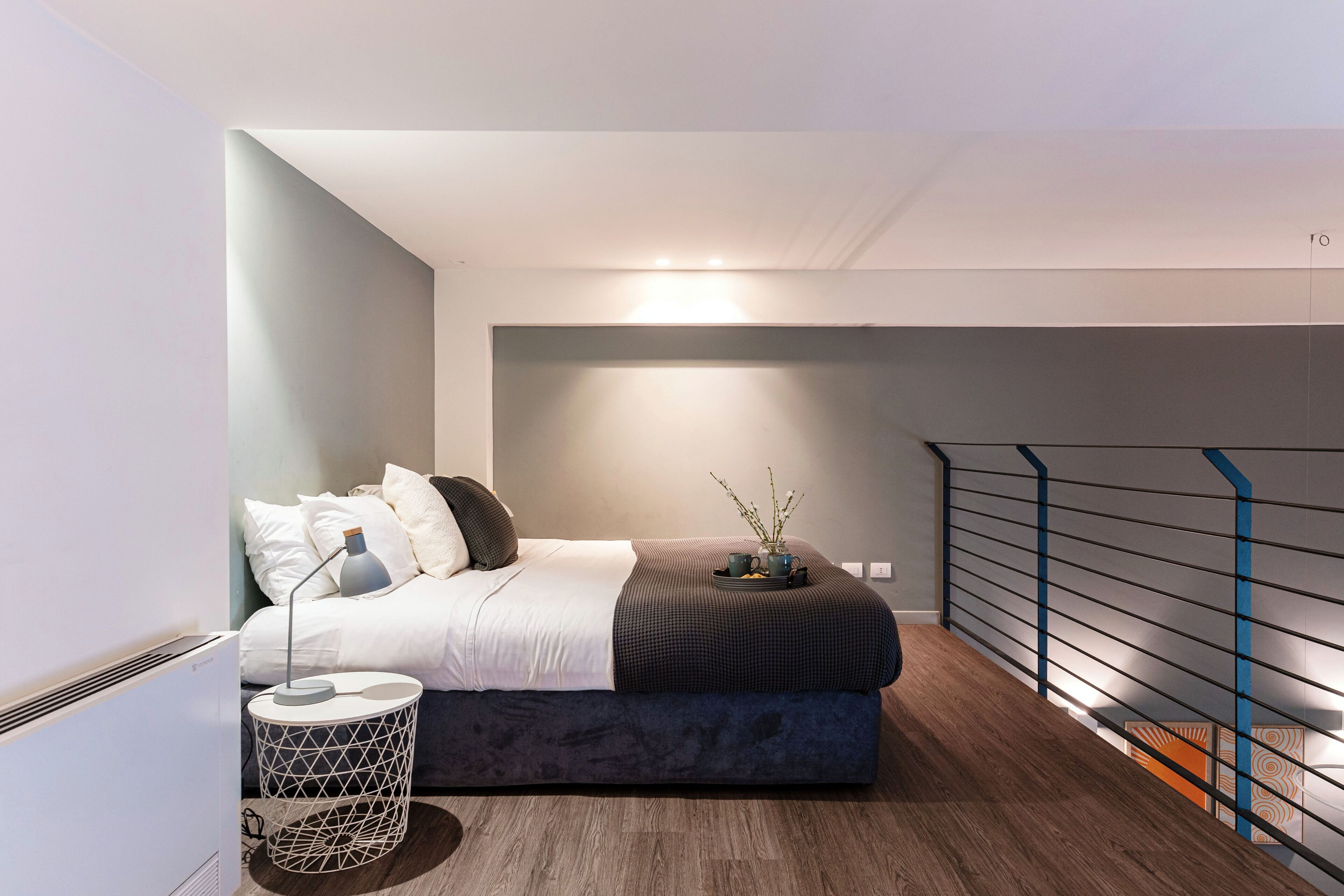 Quarto moderno com cama, mesa de cabeceira, abajur e vista para a escadaria.