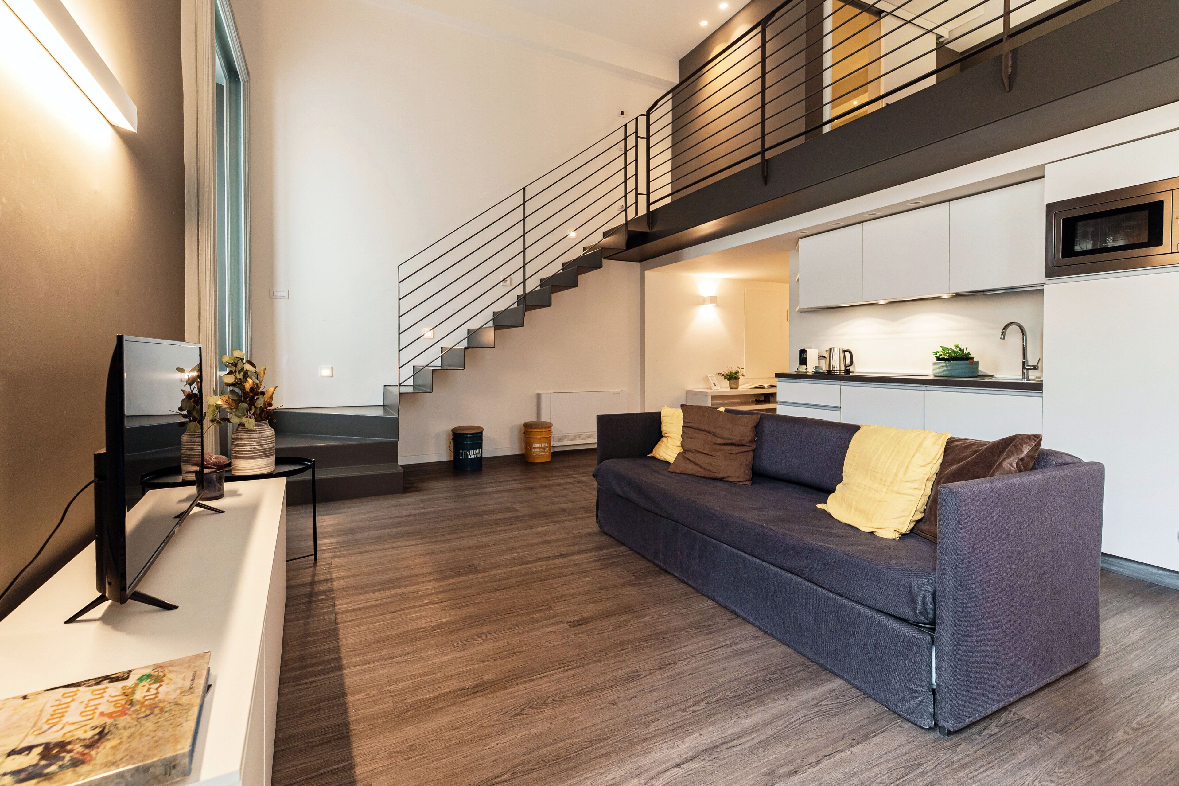 A modern living room with a sofa, a flat-screen TV, a staircase, and a kitchen area.