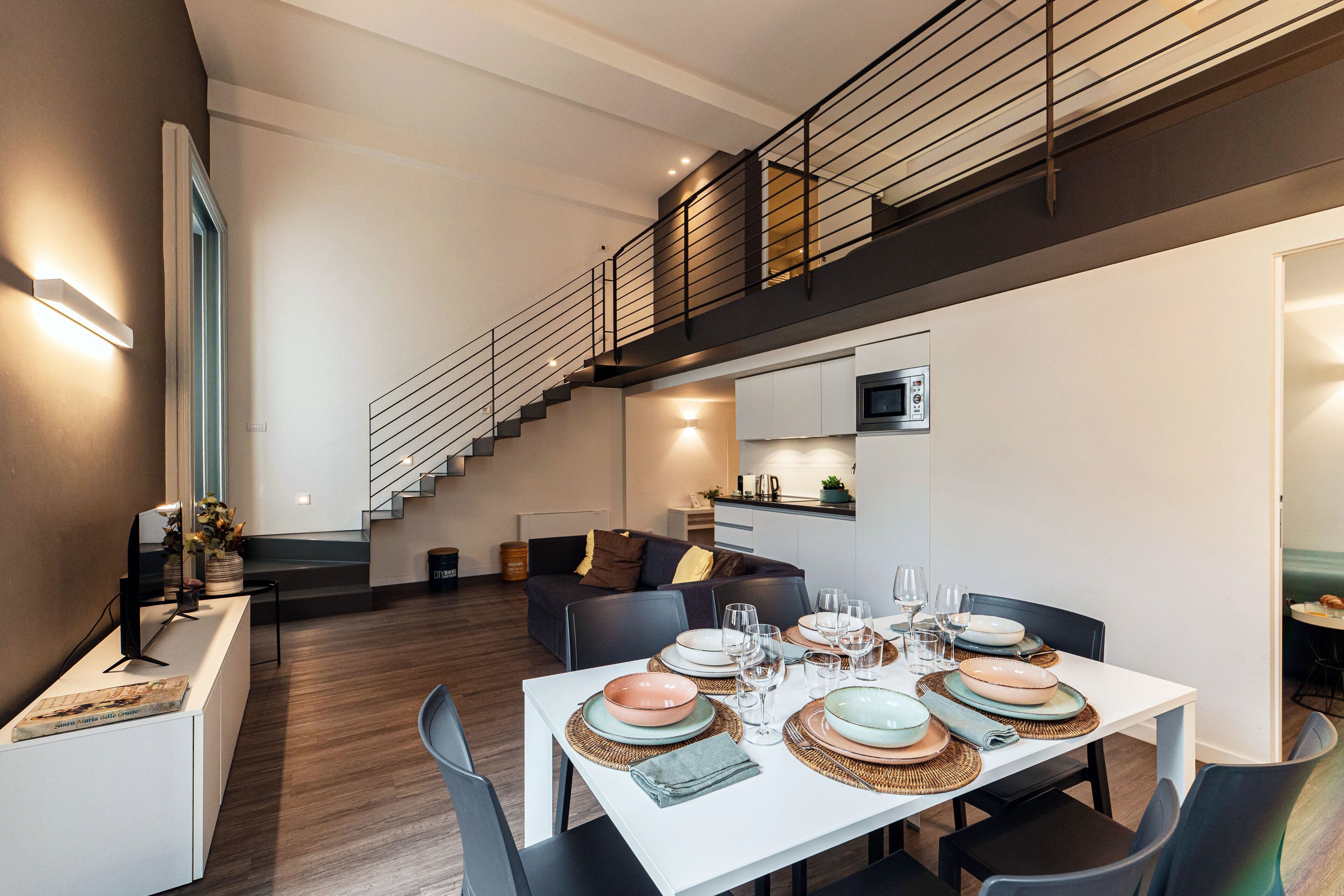 A modern dining area with a white table set for six, a staircase, and a kitchen area with a microwave.