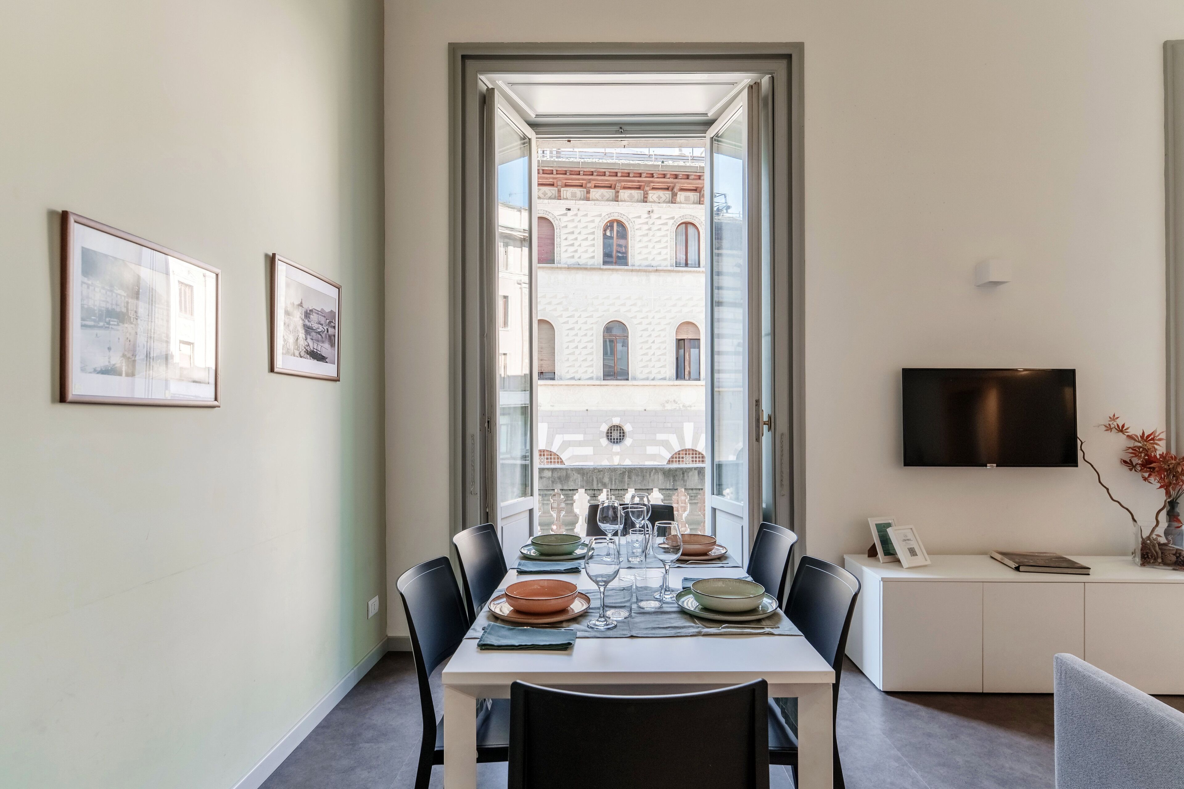 A dining area with a table set for six, a TV mounted on the wall, and a view of a building through a glass door.