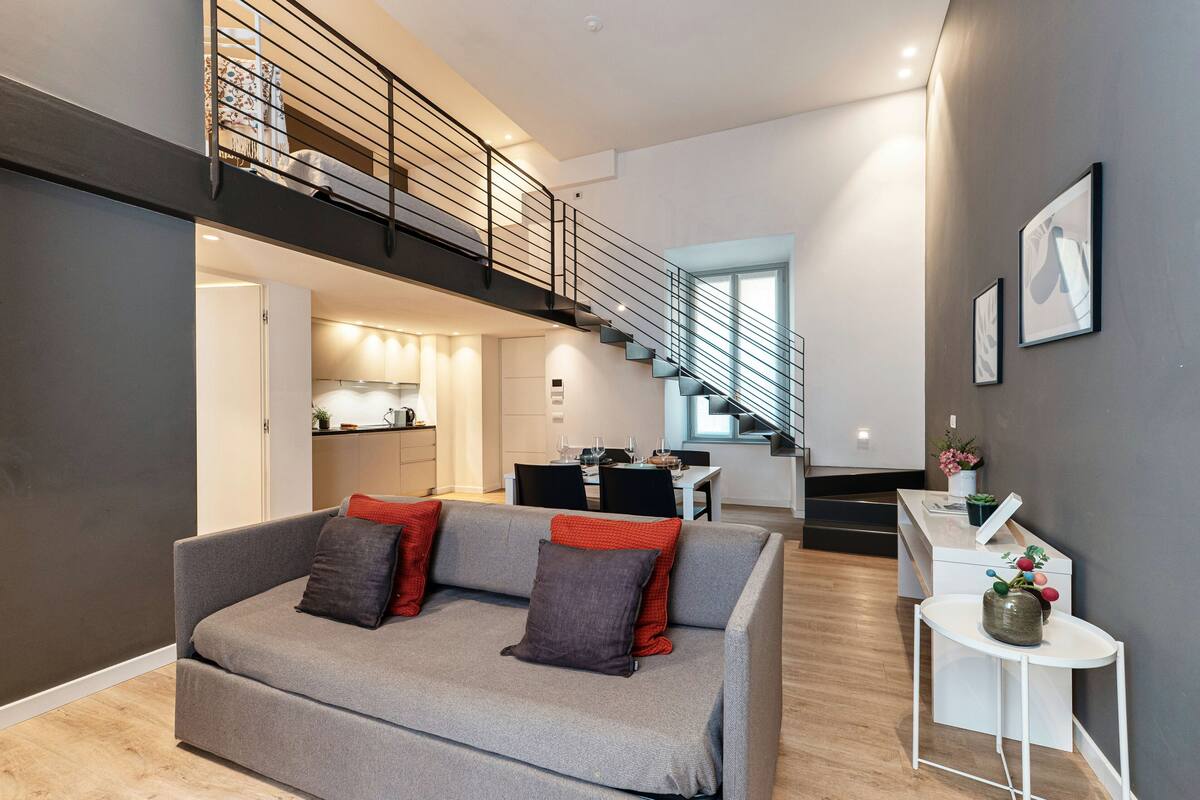 A modern living room with a grey sofa, a staircase, and a kitchen in the background.