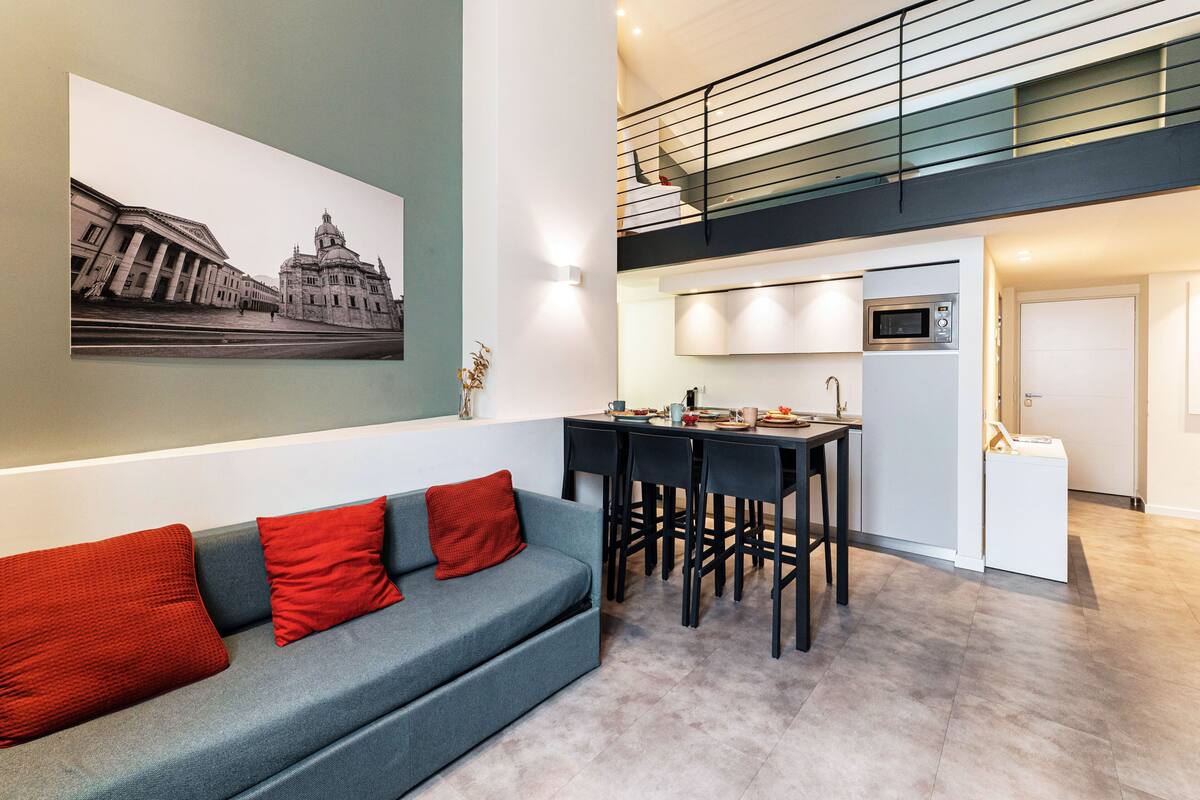 A modern interior with a grey sofa, red cushions, a dining area with chairs, a kitchen with a microwave, and a staircase.