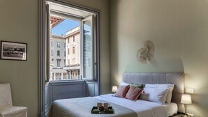Ein Hotelzimmer mit Bett, Nachttischlampen, einem Fenster mit Blick auf Gebäude, einem Sessel und einem Tablett mit Tassen.