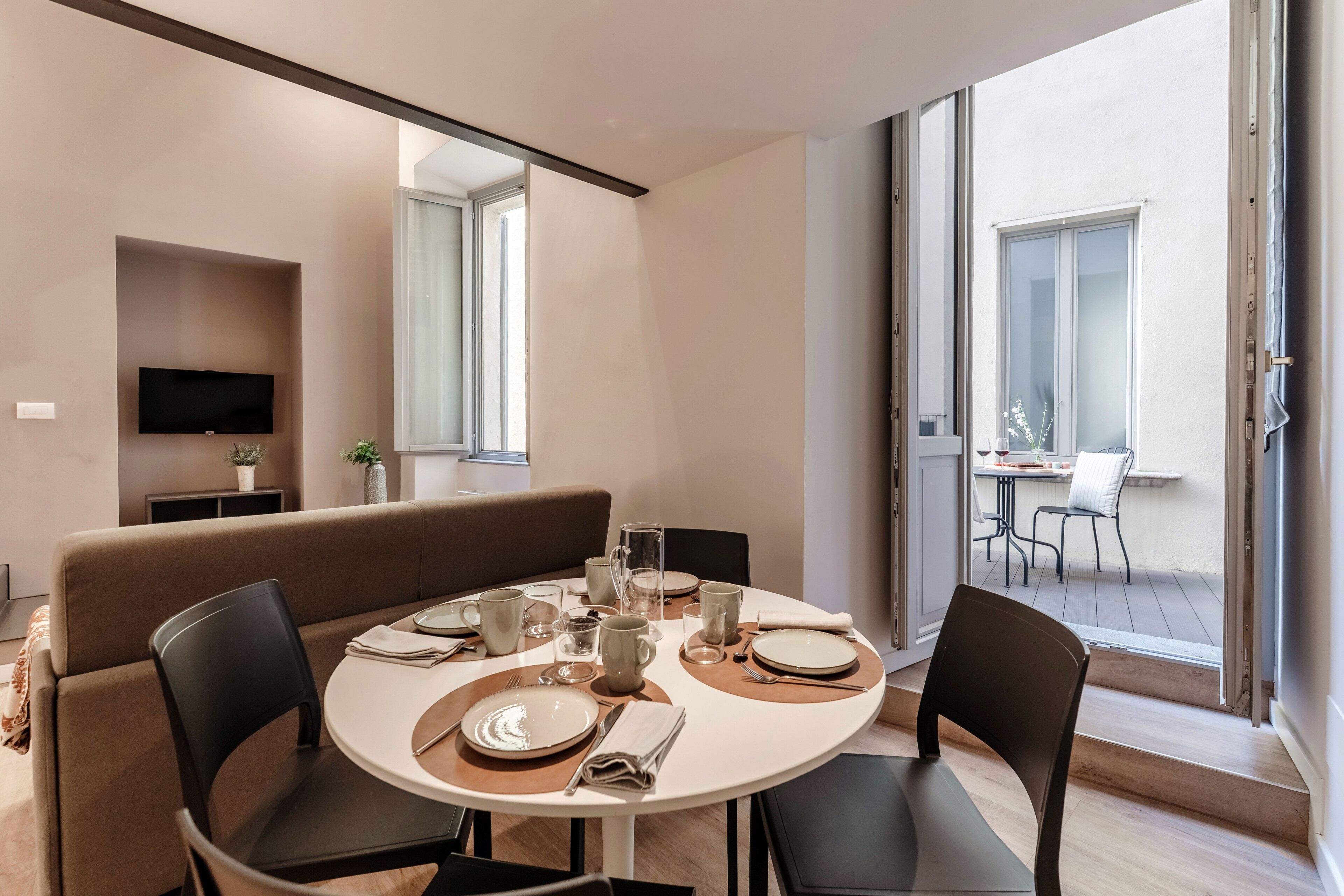 A modern dining area with a round table, chairs, and a sofa. A TV is mounted on the wall, and there is a balcony with a table and chairs.