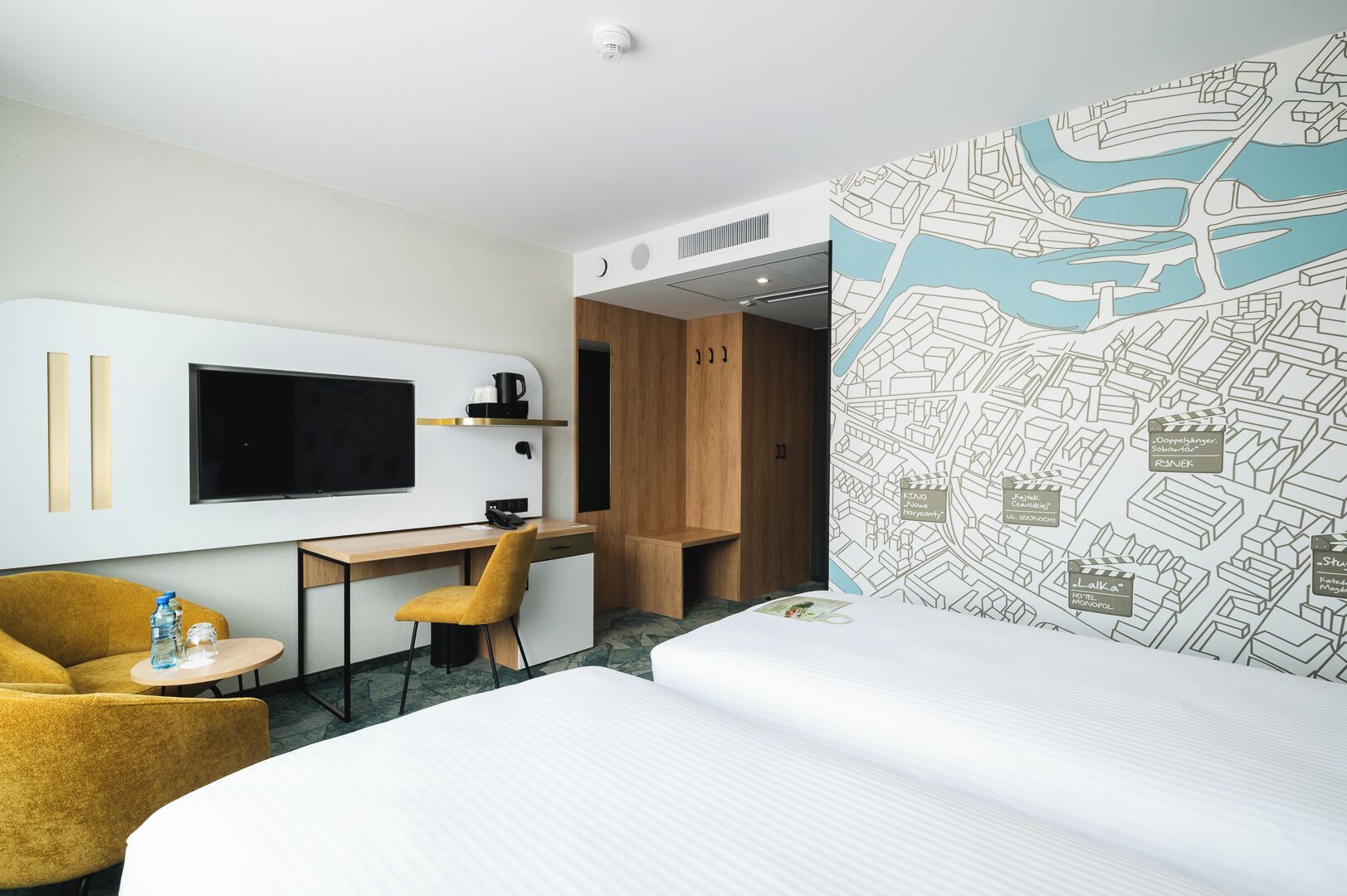 A hotel room with a bed, a desk, a chair, a TV, and a map on the wall.