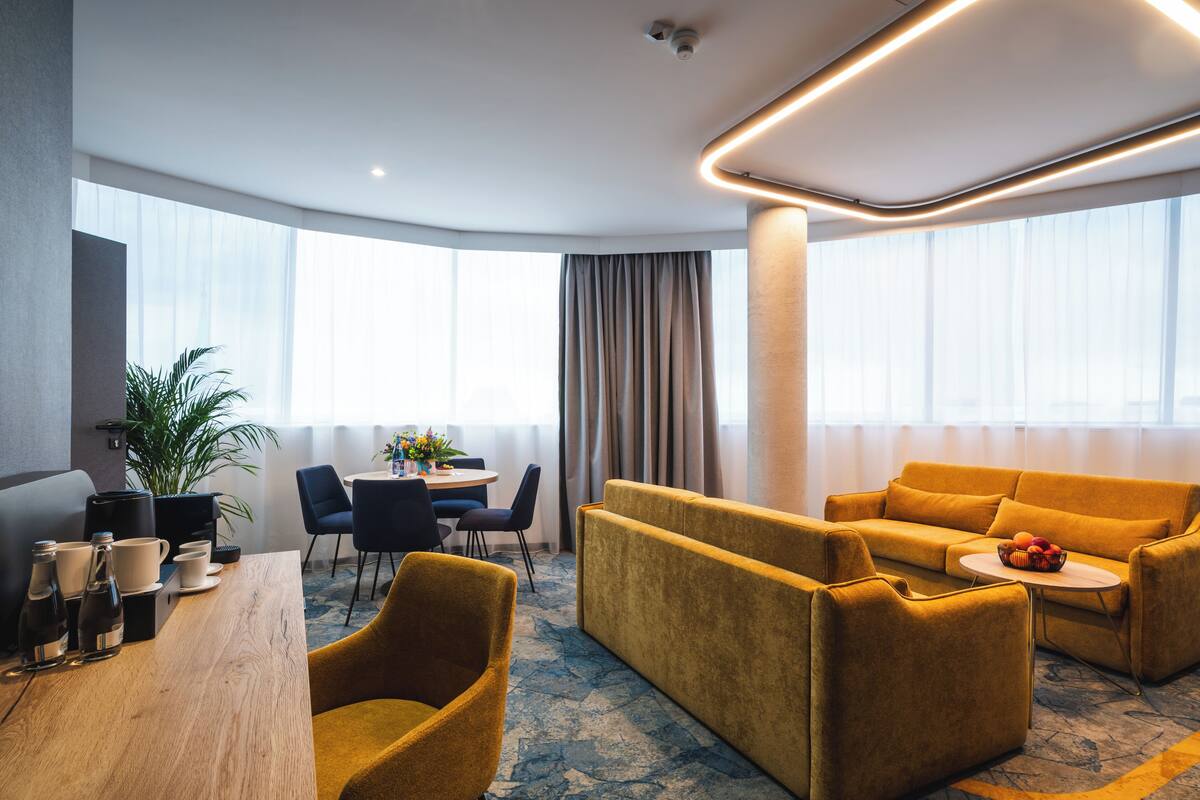 A modern hotel room with a sofa, dining table, chairs, and a potted plant.