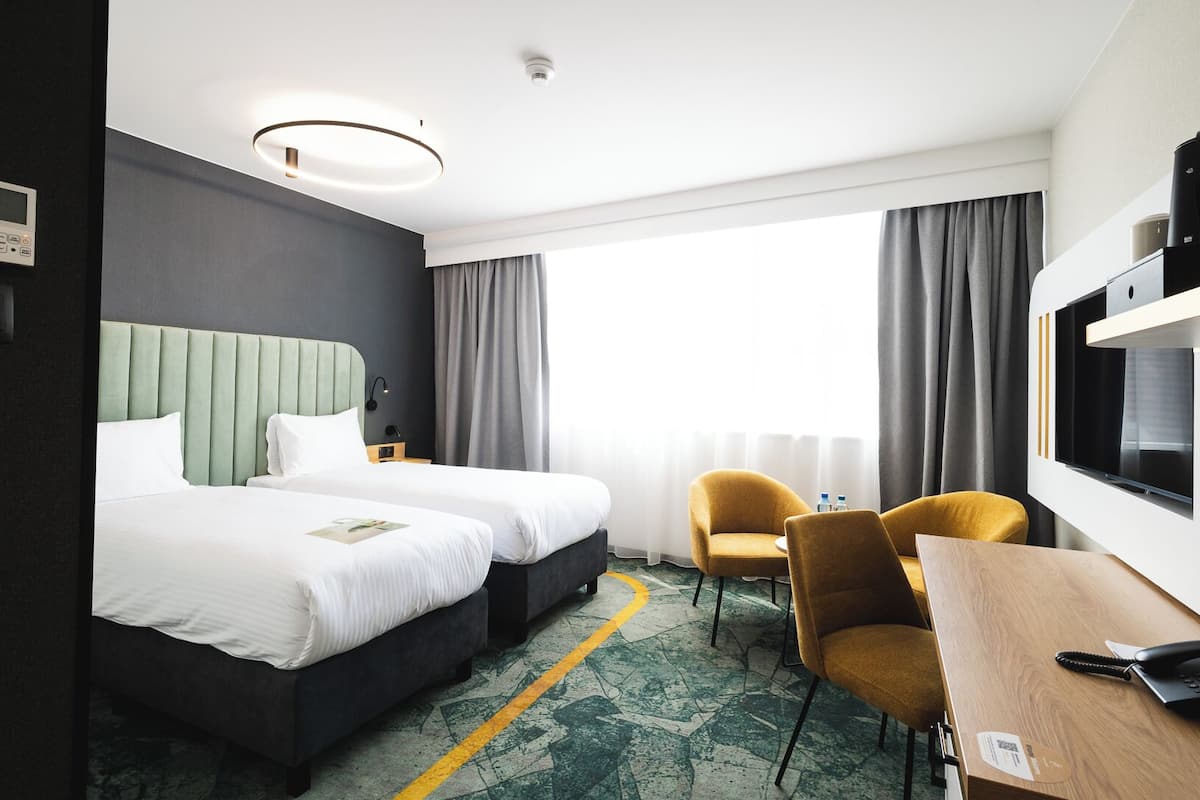 A hotel room with two beds, a desk, chairs, a TV, and a large window with curtains.