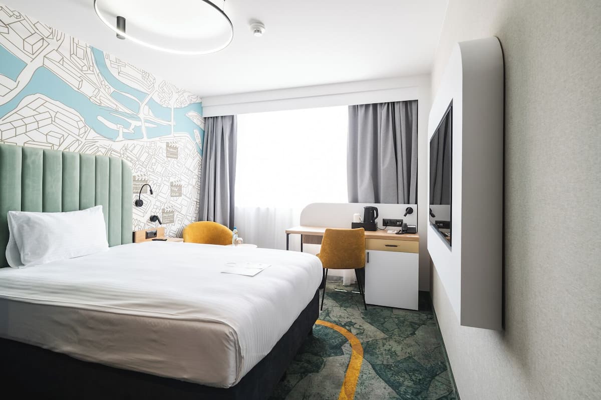 A hotel room with a large bed, a desk with a chair, a desk lamp, a coffee maker, and a large window with curtains.