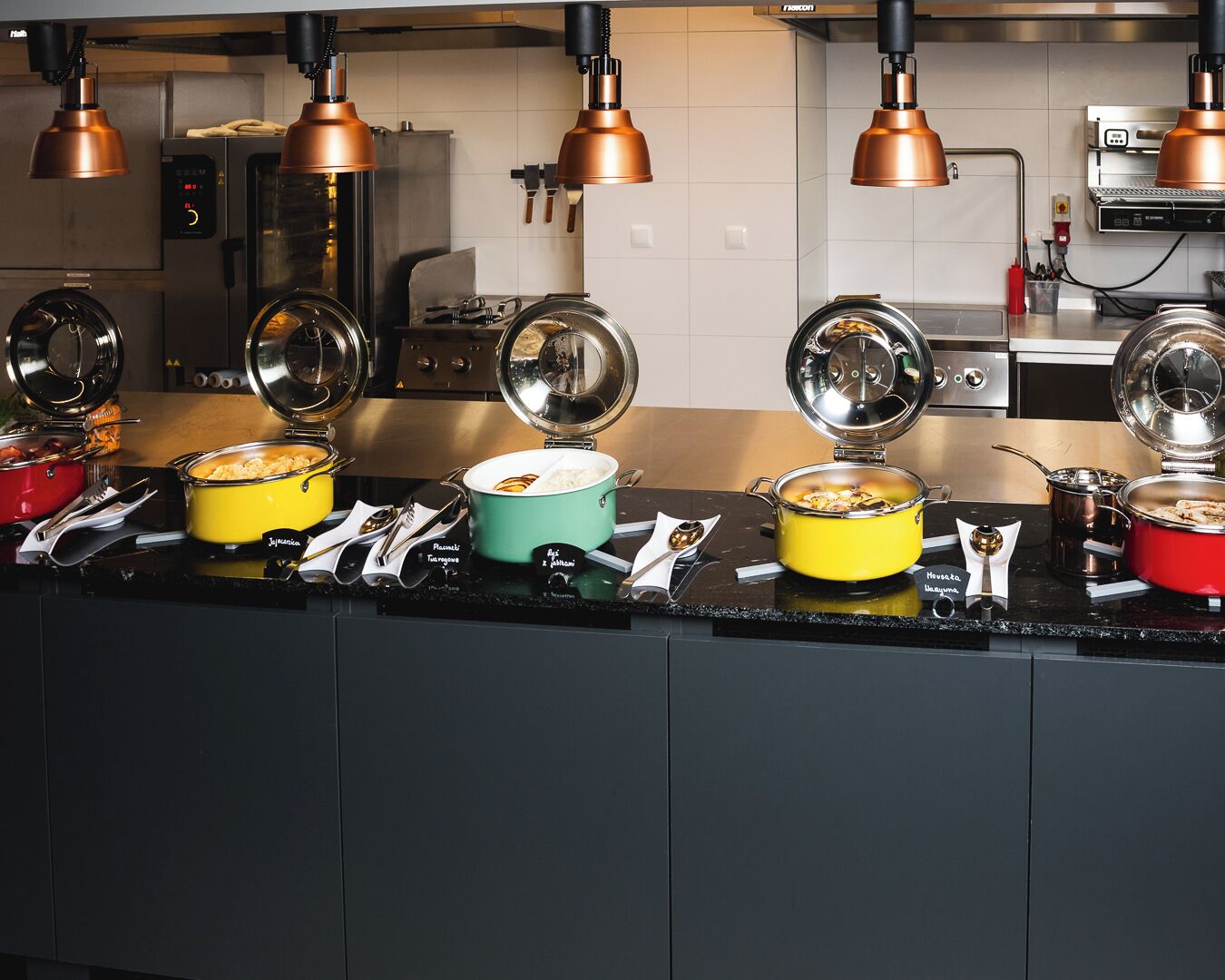 Una cocina con varias ollas sobre la hornilla, iluminación de cobre colgante y un electrodoméstico moderno al fondo.