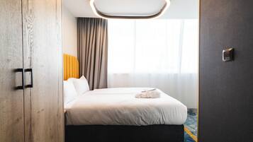 Habitación de hotel con cama, cabecero, mesita de noche, lámpara de pared y ventana con cortinas.