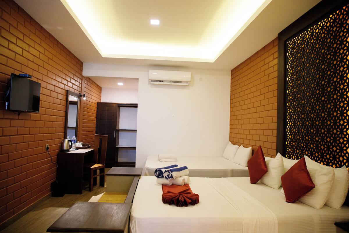 Habitación de hotel con pared de ladrillo, un televisor de pantalla plana, un escritorio con silla, cama con ropa de cama blanca y almohadas rojas, y un panel decorativo en la pared.