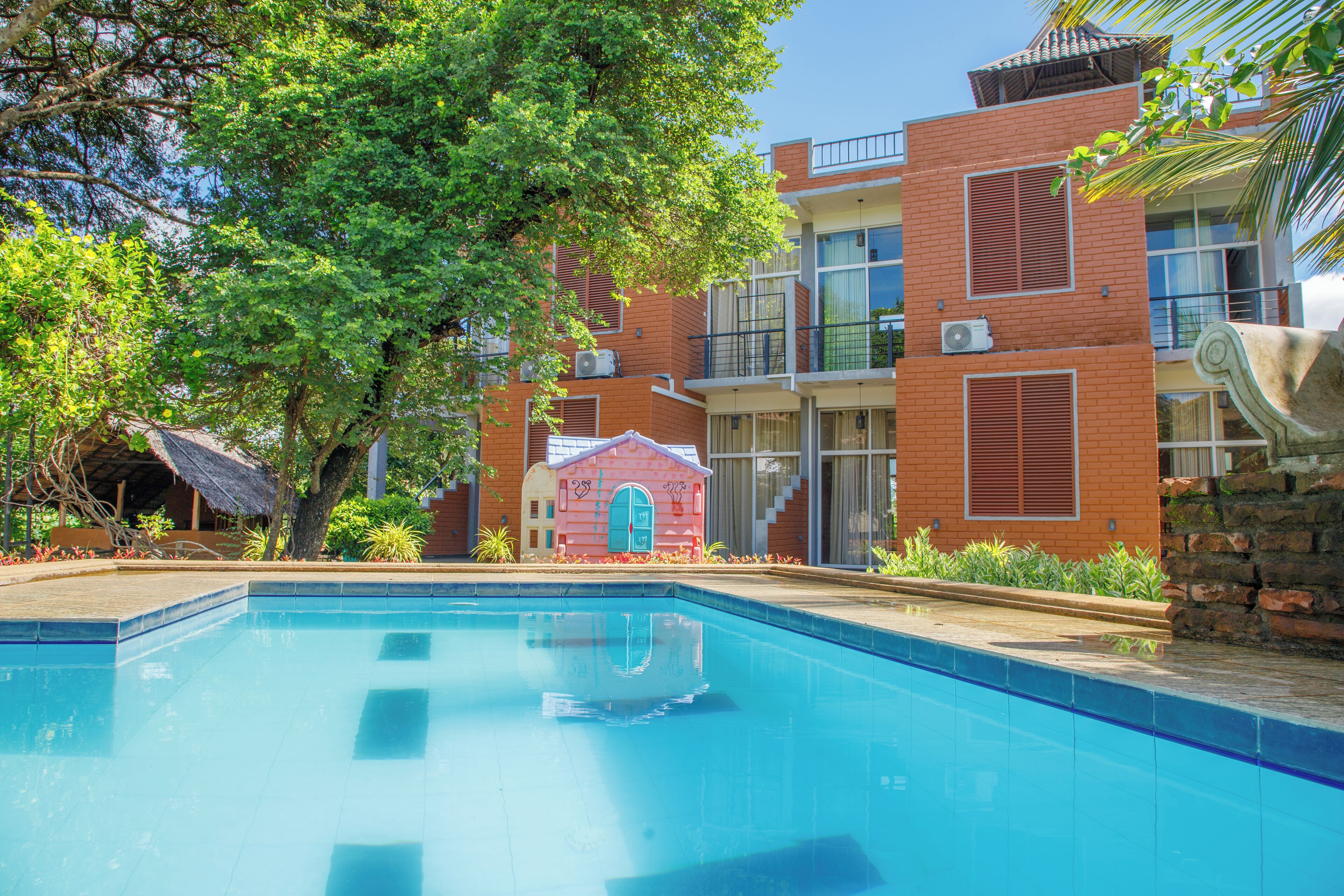 A swimming pool with a clear blue water surface, surrounded by a brick building with balconies and a small pink structure with a blue door.