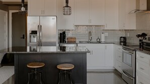 A modern kitchen with white cabinets, a stainless steel refrigerator, a built-in oven, and a large island with stools. - Modern Industrial Loft for Two (Benton Harbor)