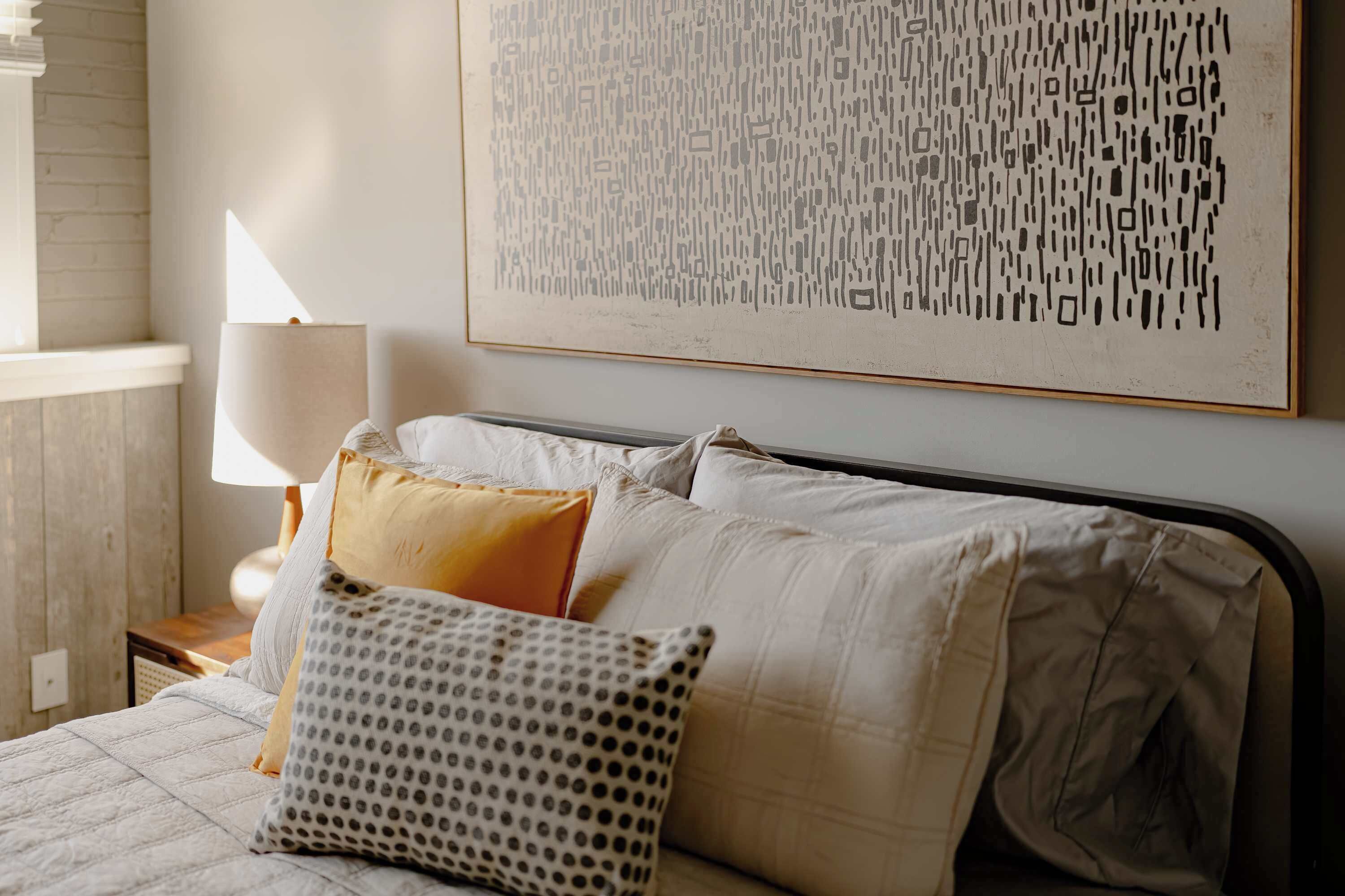 Un lit bien fait, agrémenté d’oreillers à motifs, une lampe de chevet et une grande œuvre d’art abstraite accrochée au mur.