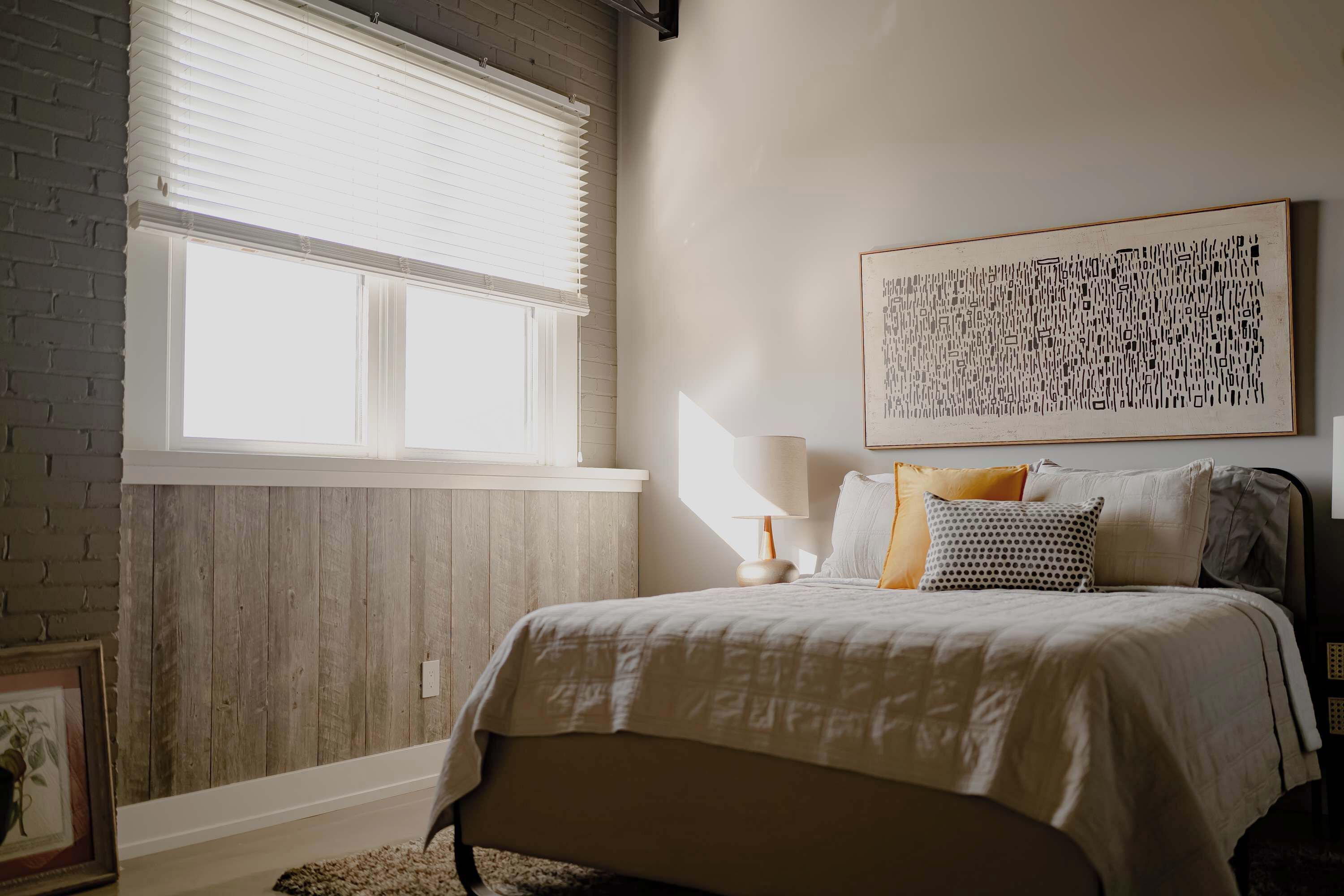 A bedroom with a bed, a window with blinds, a wall-mounted lamp, and a framed picture.