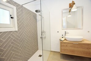 Un baño moderno con salpicadero de baldosas en espiga, ducha con mampara de vidrio, tocador de madera con lavabo blanco e iluminación colgante en un espejo.