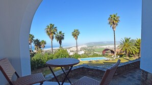 A balcony with wicker chairs and a small table, overlooking a pool and a view of the ocean and distant hills.