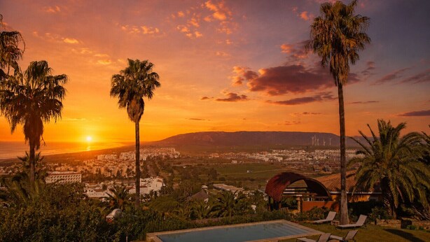 A pool with lounge chairs, palm trees, and a view of a cityscape at sunset.