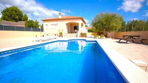 Une piscine avec un bord carrelé, un espace patio doté d’une table de pique-nique et un bâtiment avec un toit recouvert de tuiles.