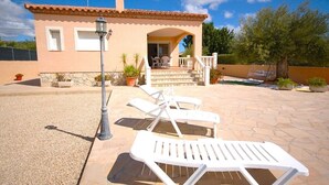 Une maison dotée d’un patio aménagé avec des chaises longues blanches et une table, ainsi qu’une lampe et des plantes en pot.