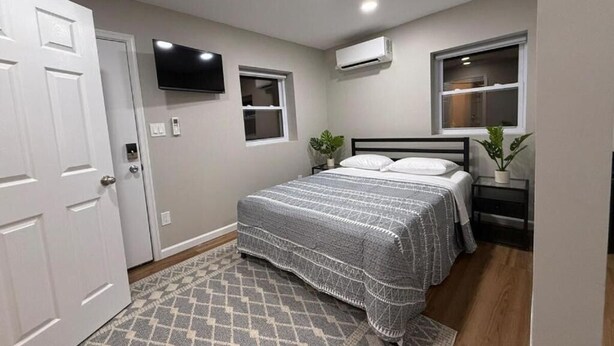 A bedroom with a bed, a TV mounted on the wall, an air conditioning unit, and a window.