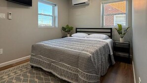 A bedroom with a bed, a TV, an air conditioner, and two windows.