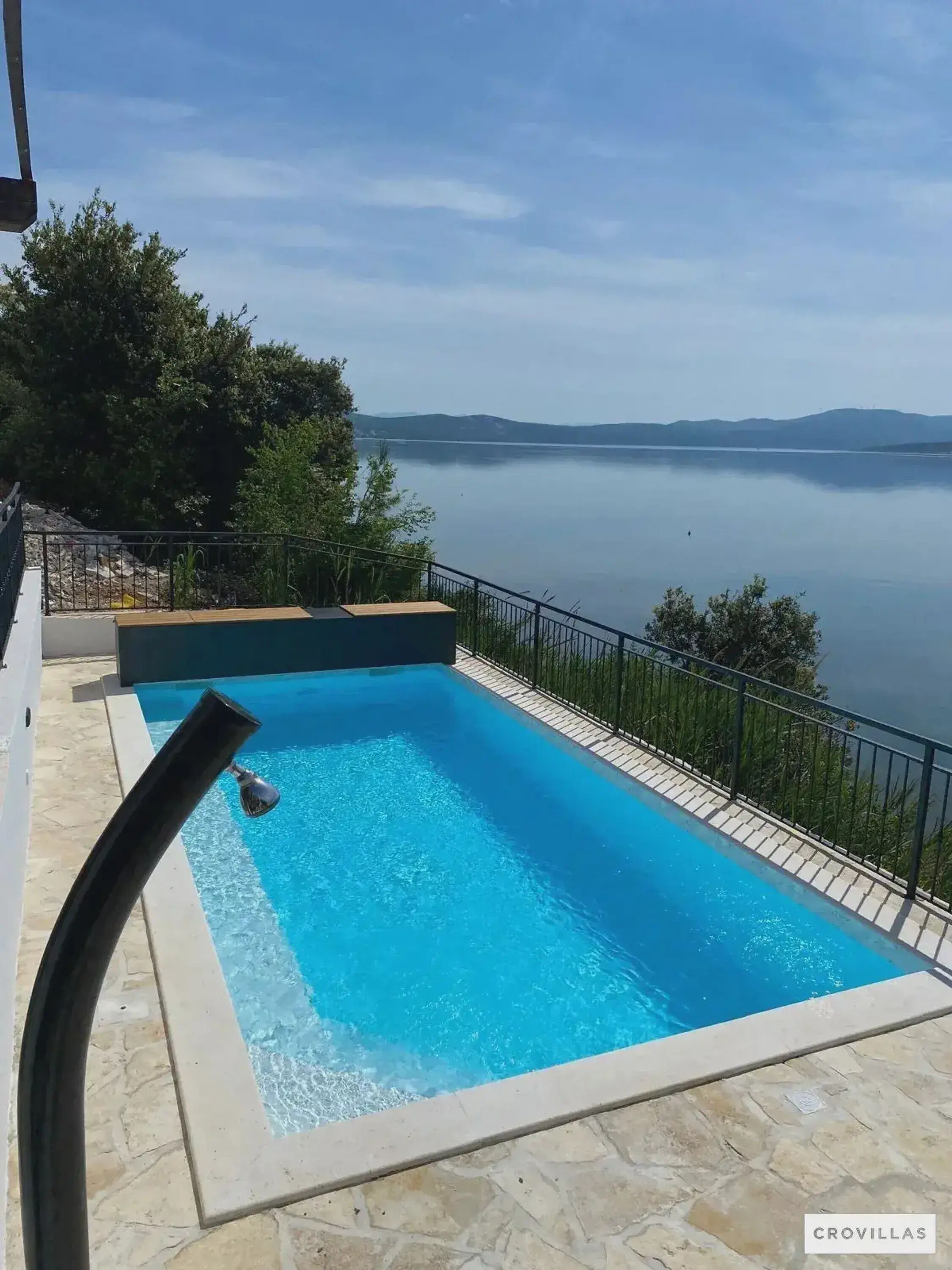 A rectangular swimming pool with clear blue water, surrounded by a stone deck and a metal railing.