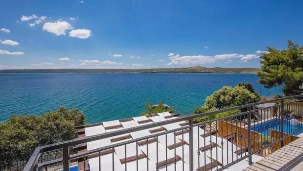Un balcón con piscina, con vista al mar azul y colinas lejanas.