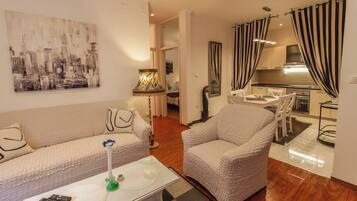A modern living room with a sofa, armchair, and coffee table. There is a painting on the wall, a lamp, and a kitchen area visible through an open door.