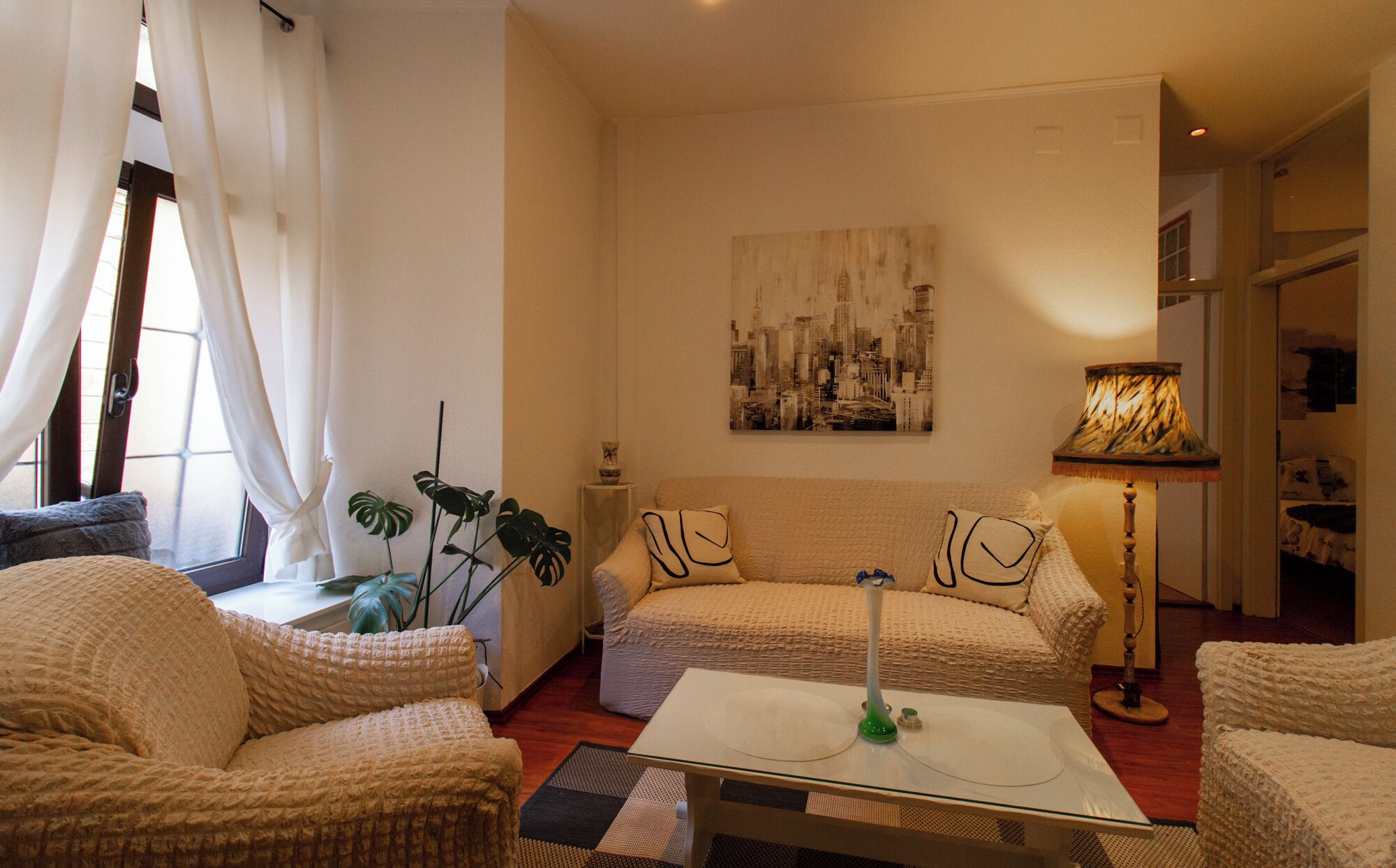 Ein gemütliches Wohnzimmer mit einer Couch, zwei Sesseln, einem Couchtisch und einem Stadtbild an der Wand.