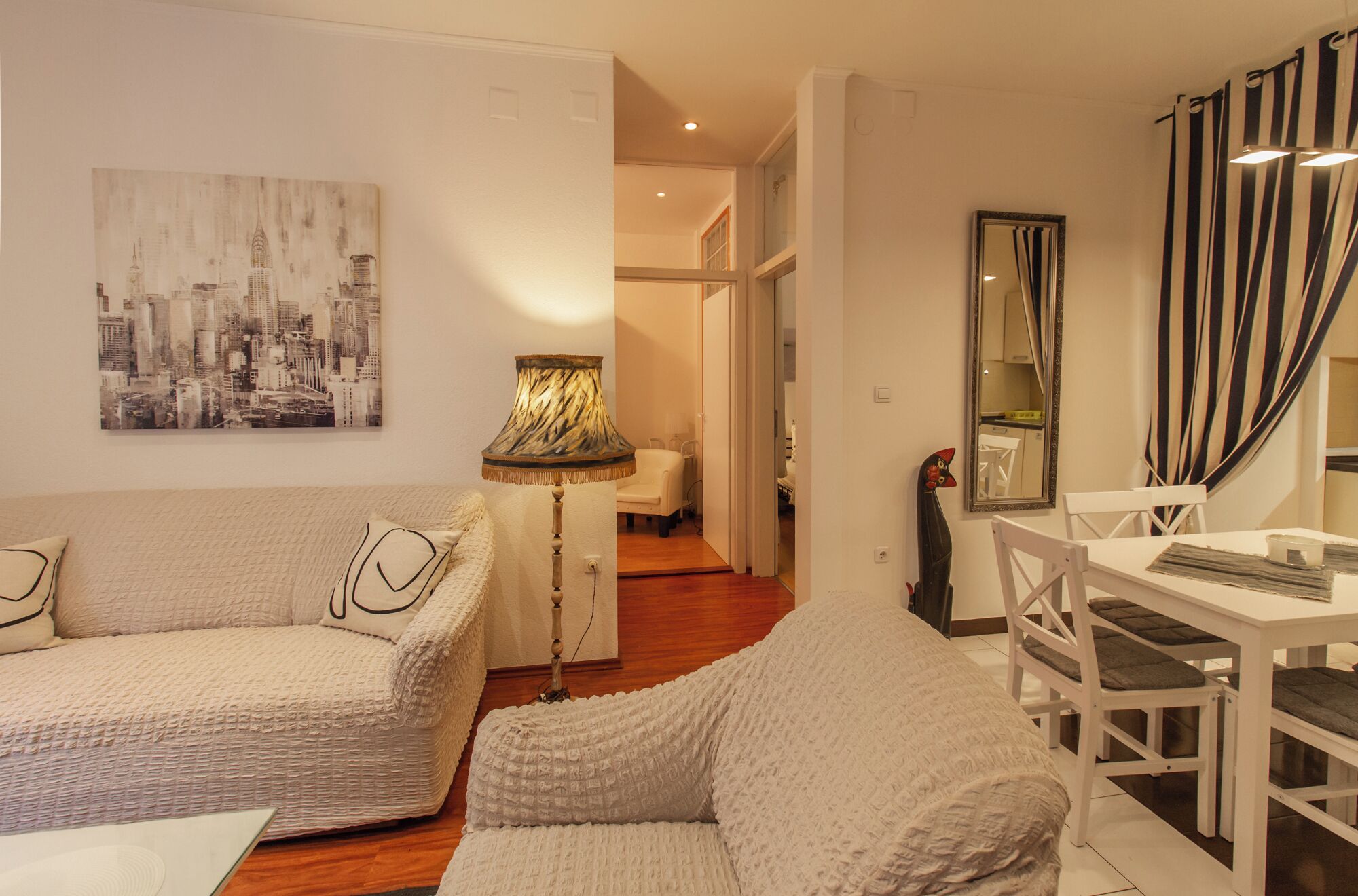 A living room with a sofa, a lamp, a dining table with chairs, and a cityscape painting on the wall.