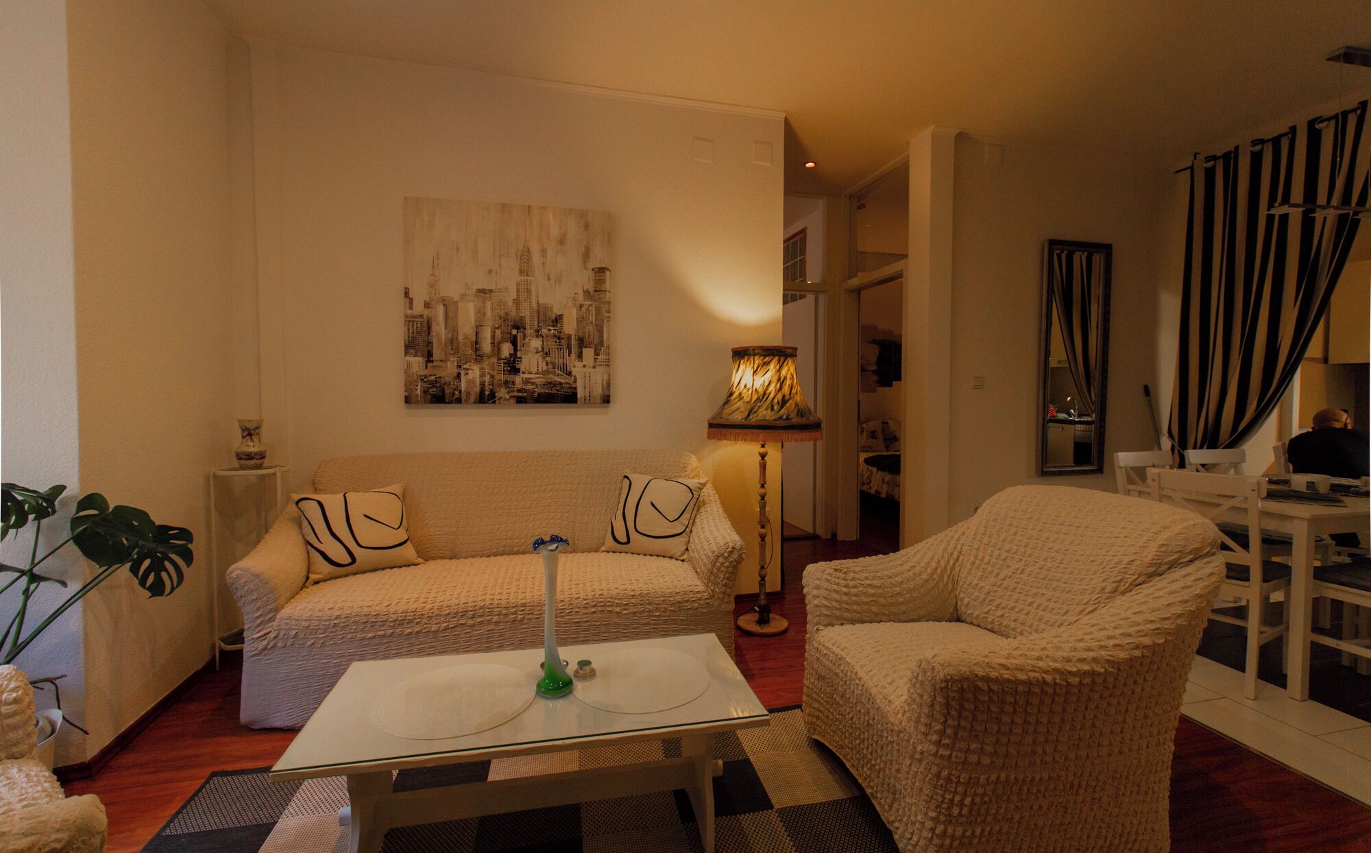 A cozy living room with a sofa, armchair, and a glass coffee table. A cityscape painting adorns the wall, and a lamp provides soft lighting.