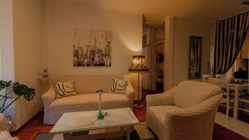A cozy living room with a sofa, armchair, and a glass coffee table. A cityscape painting adorns the wall, and a lamp provides soft lighting.