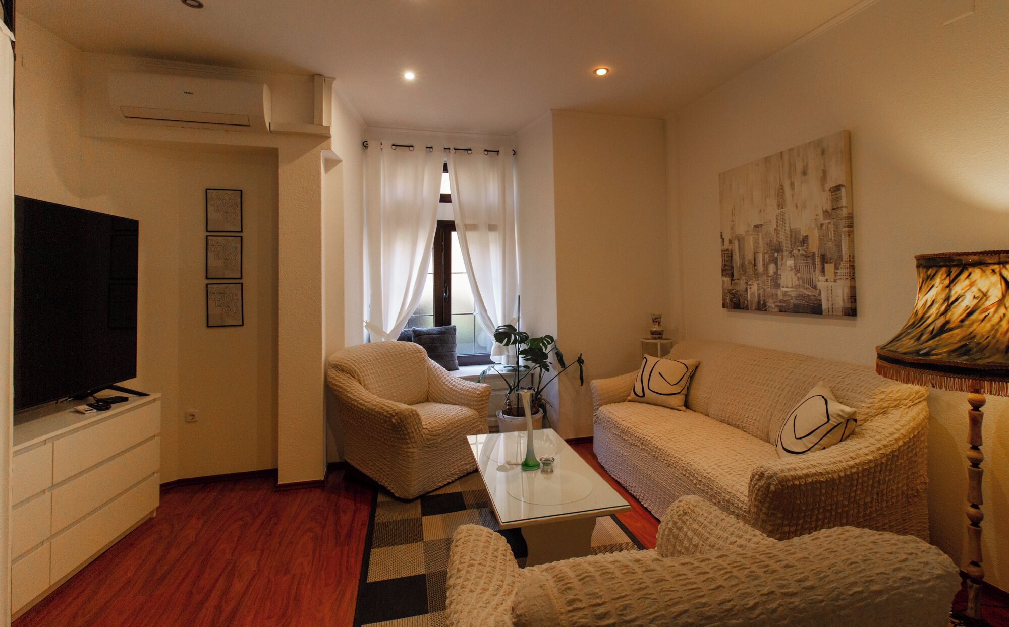 A cozy living room with a flat-screen TV, a sofa, two armchairs, a coffee table, and a cityscape painting.