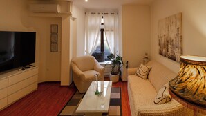 Un soggiorno moderno con una televisione a schermo piatto, un divano color beige, una poltrona coordinata, un tavolino da caffè e un'ampia finestra con tende leggere.