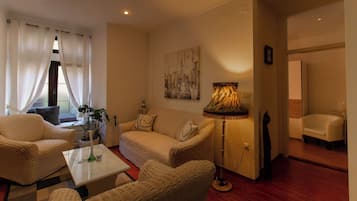 A cozy living room with a sofa, two armchairs, a coffee table, and a floor lamp. There is a window with sheer curtains, a cityscape painting on the wall, and a door leading to another room.