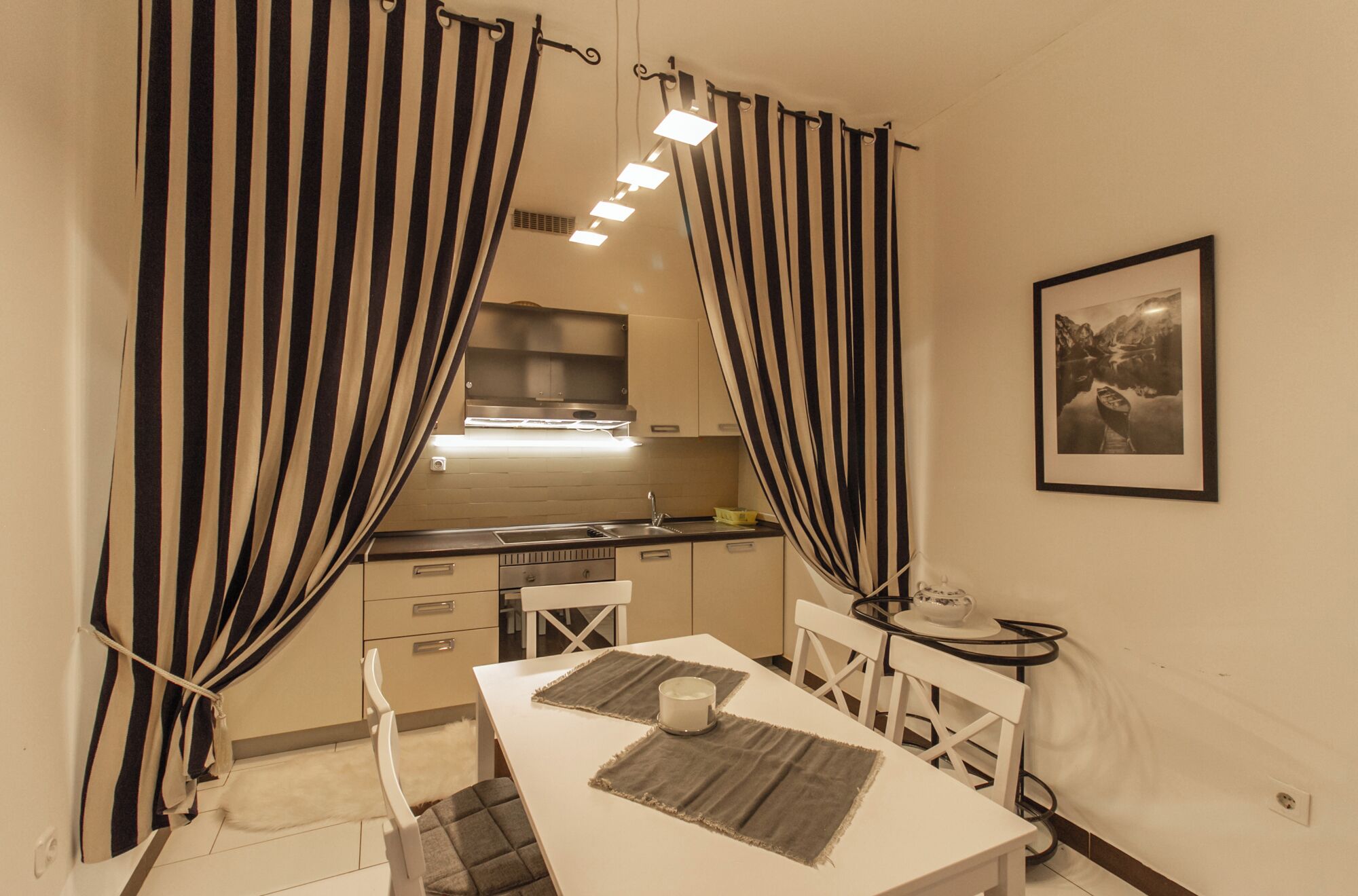 A compact kitchen with a modern stove, a sink, and a small countertop. There is a dining table with two chairs and a small round table with a teapot. The room has black and white striped curtains and a framed picture on the wall.
