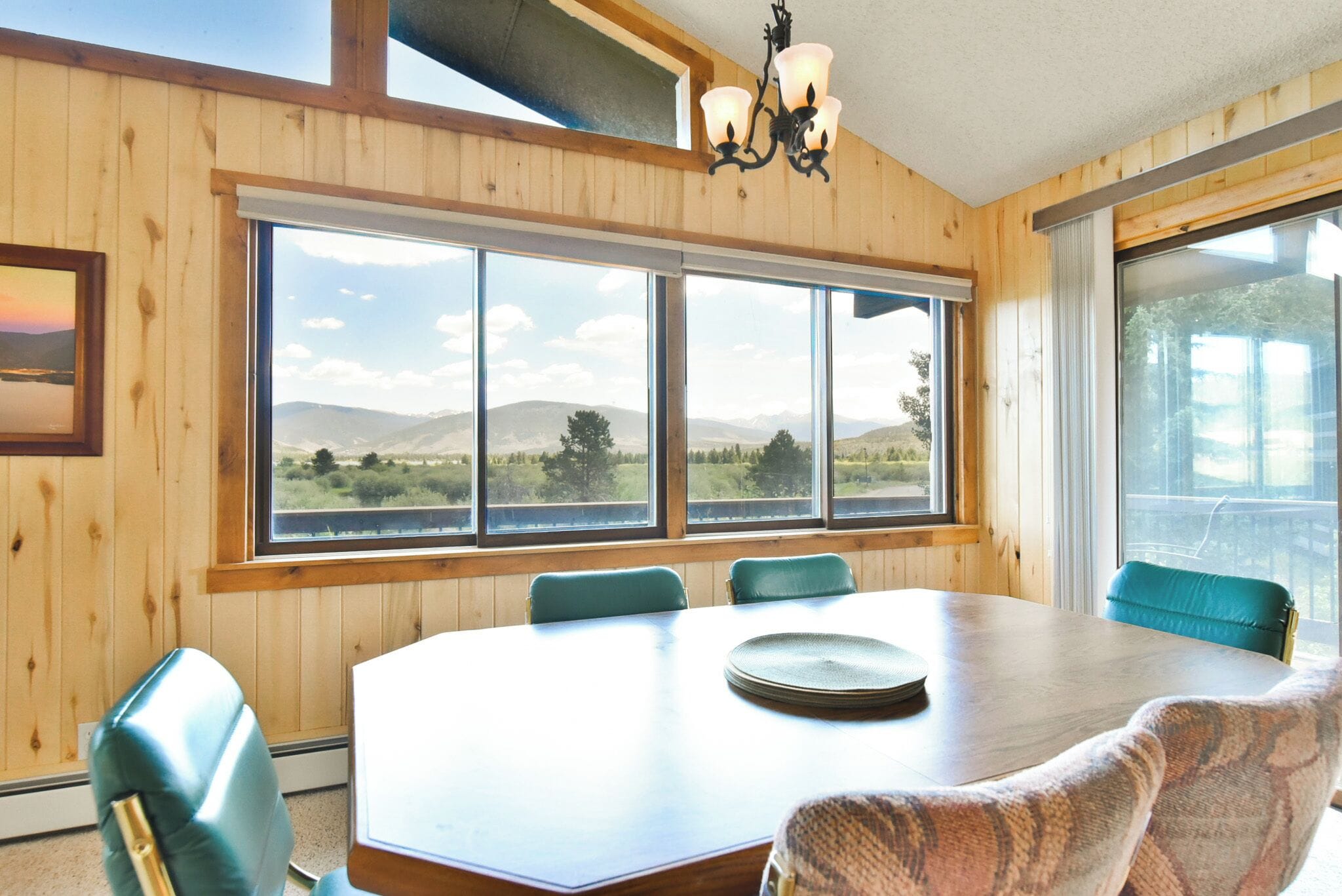 Una habitación con paneles de madera y un amplio ventanal que ofrece vistas a montañas y un lago. Hay una mesa redonda con sillas y una araña.