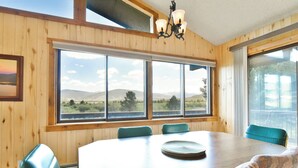 Ein Zimmer mit Holzwänden und einem großen Fenster mit Blick auf Berge und einen See. Ein runder Tisch mit Stühlen und eine Deckenleuchte sind vorhanden.