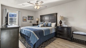 Quarto com cama, penteadeira, mesa de cabeceira, abajur e ventilador de teto.