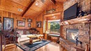 A cozy cabin interior with a wooden ceiling, a stone fireplace, a television, and framed pictures on the walls. - The Cabin at Rock Canyon (Branson)