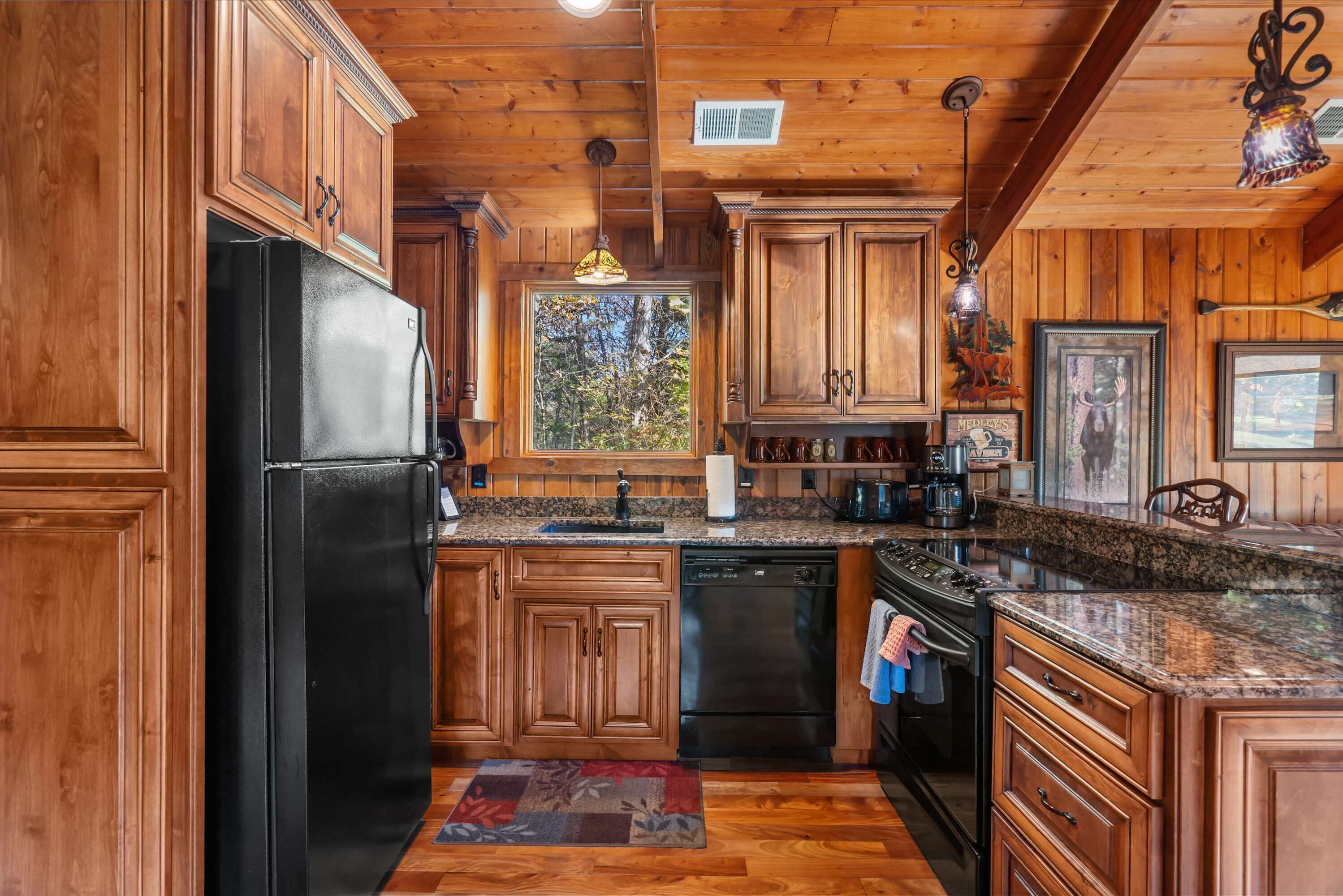 A wooden cabin kitchen with a black refrigerator, granite countertops, wooden cabinets, and a window with a view of trees.