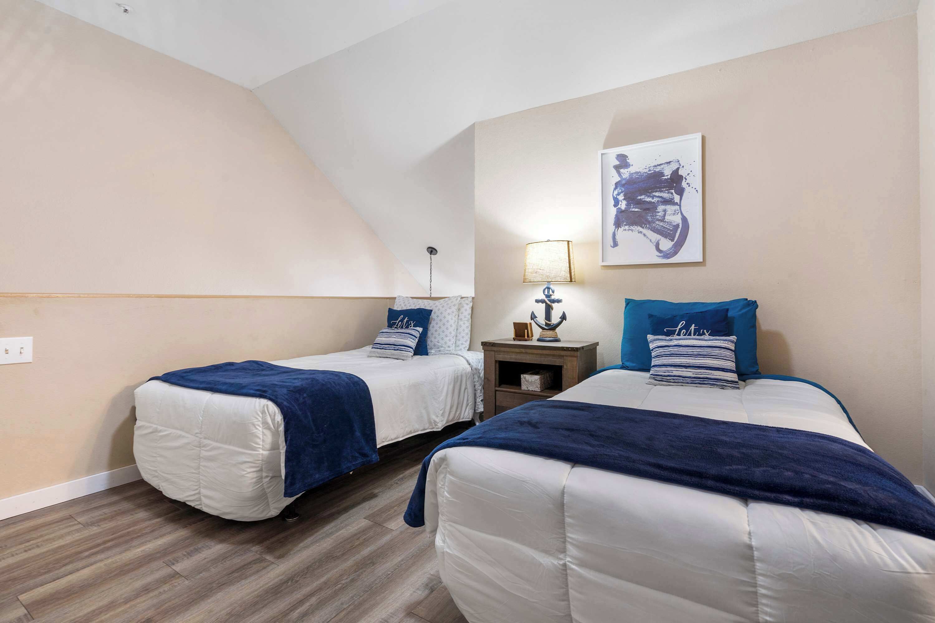A bedroom with two beds, each with a blue headboard and a white mattress. There is a nightstand with a lamp and a decorative item between the beds. A painting is on the wall above the beds.