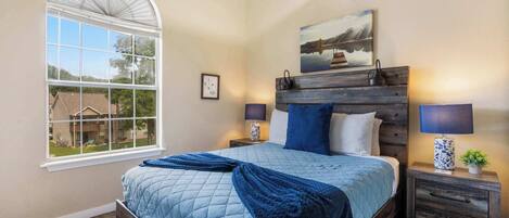 Een slaapkamer met een groot raam, een bed met een blauwe quilt, twee nachtkastjes met lampen en een schilderij aan de muur.