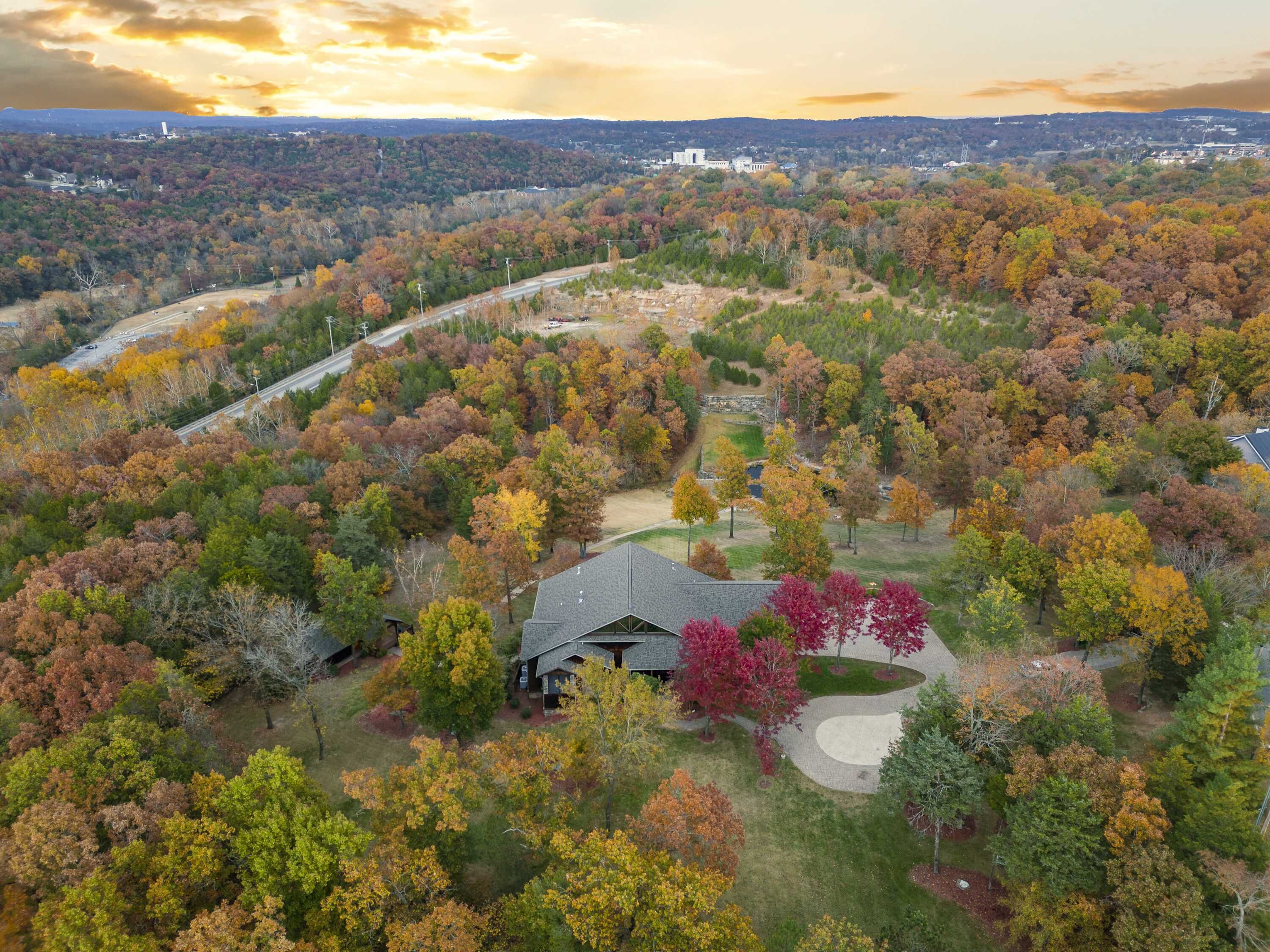 An aerial view of a residential area with a house surrounded by autumn-colored trees, a winding driveway, and a road in the background.