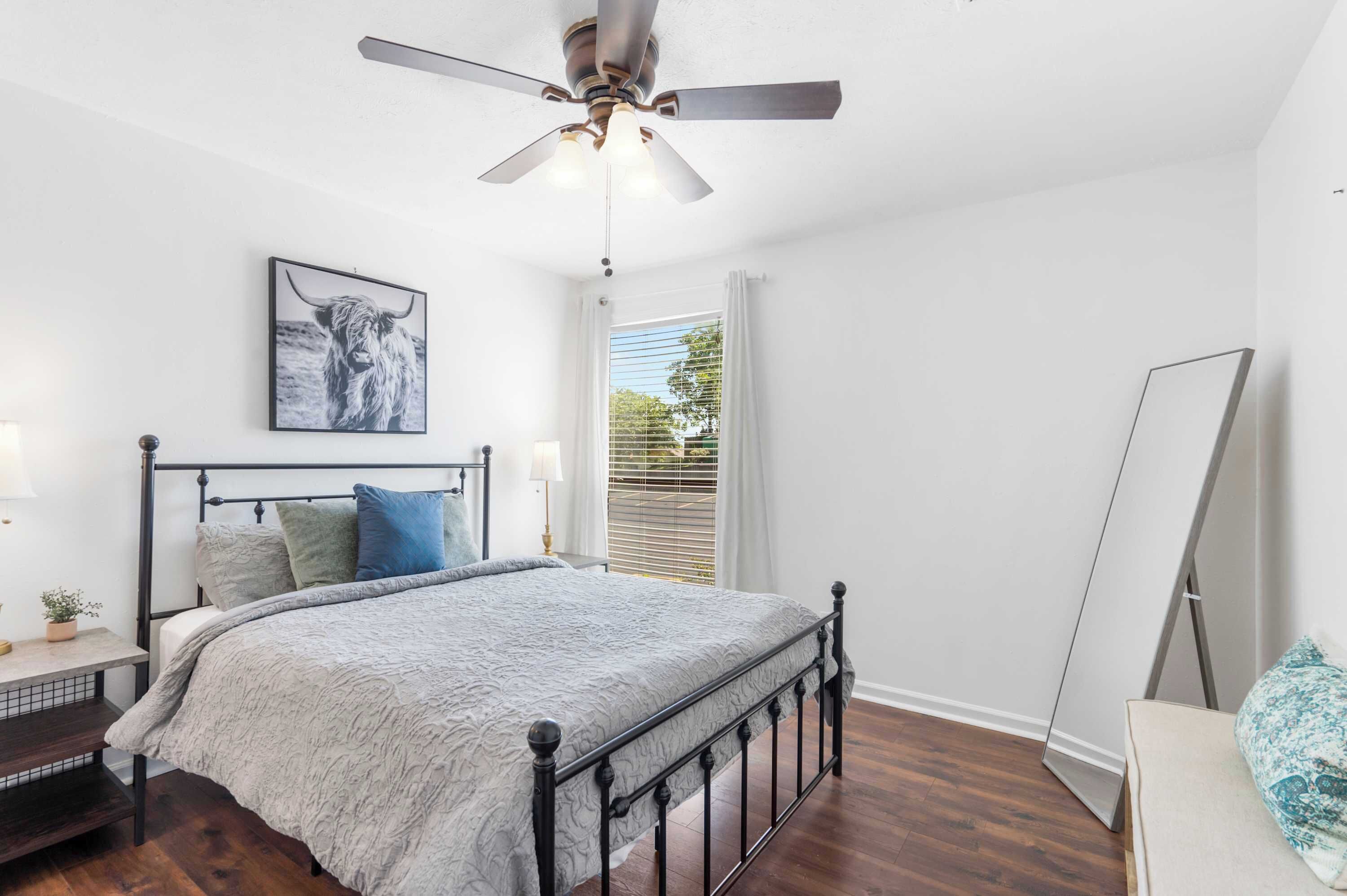 A bedroom with a bed, a ceiling fan, a window with blinds, and a framed picture on the wall.